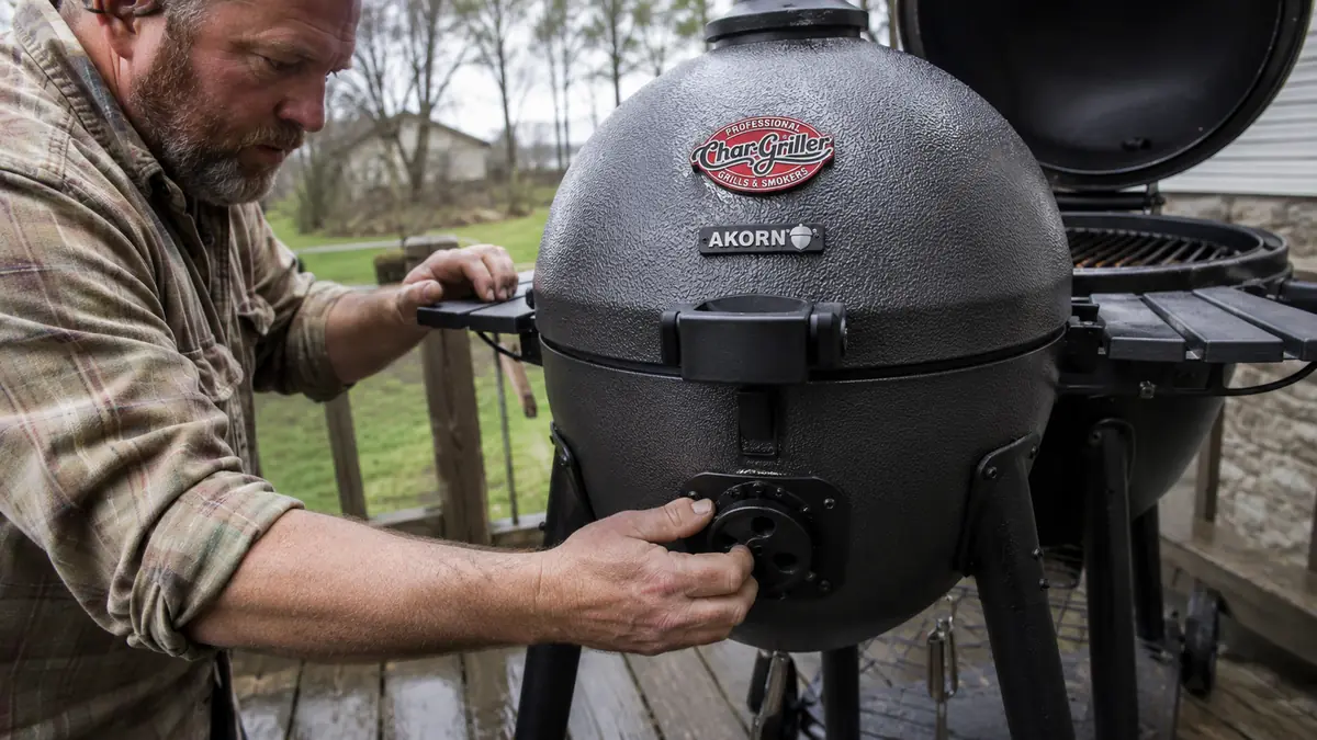 Hand adjusting the bottom vent damper on the Char-Griller AKORN while smoke curls from the top vent