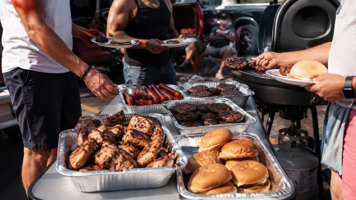 A finished tailgate spread of burgers, hot dogs, and chicken on a folding table with people reaching for plates